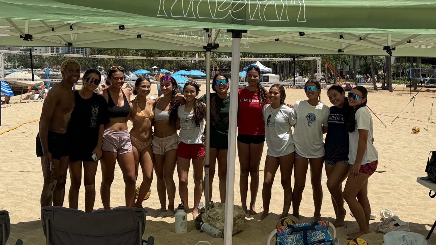 Beach volleyball players smiling together beneath a green Team Hawaiʻi Beach tent beside the courts
